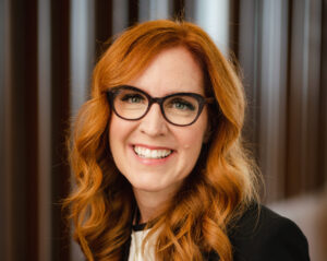 Professor RonNell Andersen Jones, a white woman with long, wavy red hair wearing glasses and a black blazer