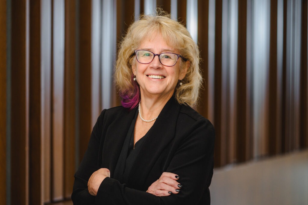Professor Jensie Anderson, an older white woman with blonde hair and glasses wearing a black blazer and pearl necklace