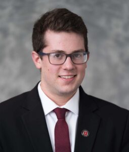 John McIntyre, a young white man with dark-brown hair and glasses wearing a black suit jacket and maroon tie