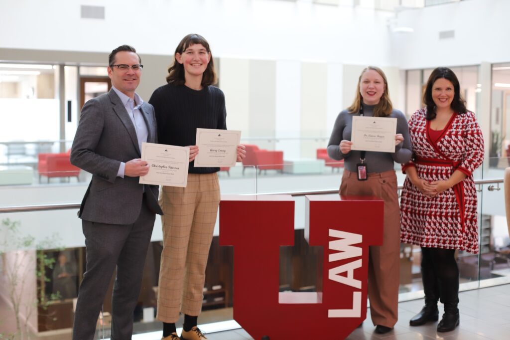 Professor Chris Peterson, left, Avery Emery, Dr. Caisa Royer, and Dean Elizabeth Kronk Warner stand near the red U Law emblem in the law school building