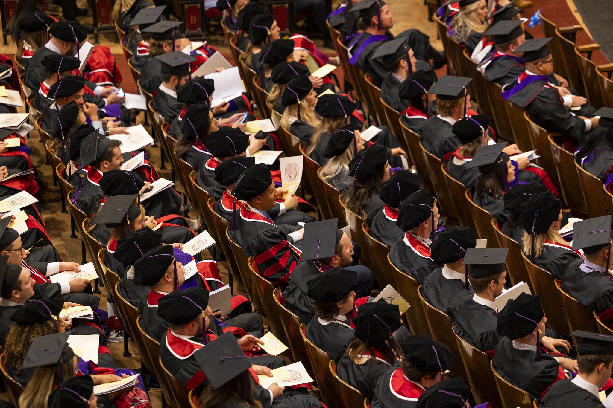 A group of graduating students sitting in rows of seats in an auditorium, all wearing graduation gowns and mortarboard hats.