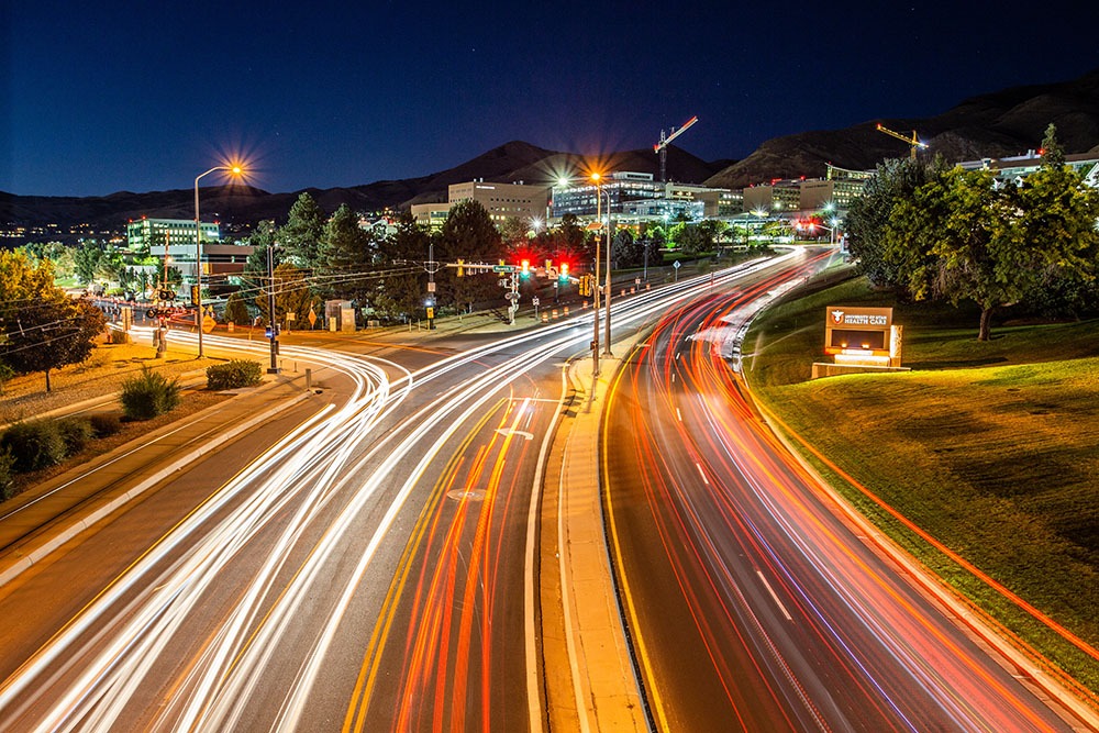 A view of the street near the University of Utah, Mario Capecchi Drive, at night with neon light streams and a cityscape in the background