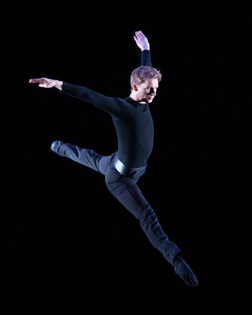 Alex McFarlin, a young white man with light-brown hair wearing a black leotard and form-fitting dance pants, leaps in the air in front of a black background