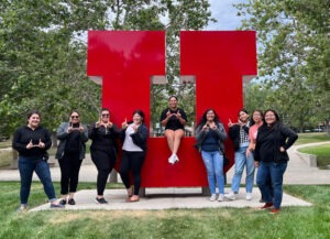 A group of eight Native women who participated in the LAILAC program make the "U" sign with their fingers and pose around the red metal U on the University of Utah campus
