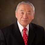 Raymond Uno, an older Japanese man with silver hair wearing a black suit with a red tie with the University of Utah logo on it