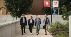 Four young law students (two men and two women) dressed in blazers and dress pants walk outside the College of Law beneath red and white flags that say "U Law" on them