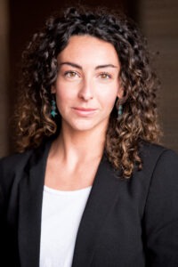 Alessandra Amato, a young Italian woman with curly, shoulder-length brown hair wearing a black blazer