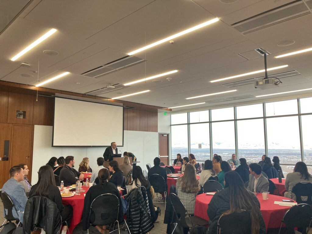 A large gathering of Native American students sitting at tables in a room and listening to a speaker standing at a lectern, with views of the Salt Lake Valley out the windows in the background
