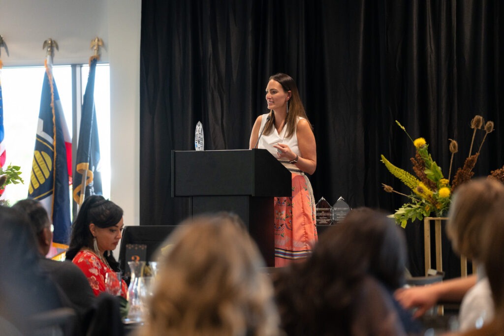 Research Professor Heather Tanana, a Native American woman with dark brown hair and olive skin wearing a white top and coral skirt, receives her award at the podium